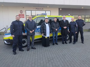 Na tle budynku wadowickiej komendy stoi oznakowany radiowóz, przed którym stoi trzech umundurowanych policjantów, ksiądz oraz trzech mężżczyzn w stroju cywilnym.