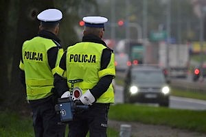 Na zdjęciu dwóch umundurowanych policjantów ruchu drogowego w kamizelkach odblaskowych z napisem Policja, stojących tyłem.  Jeden z nich trzyma w rękach policyjny miernik prędkości. W tle jadące samochody.