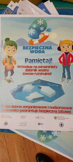 plakat Bezpieczna woda z napisem pamietaj wchodząc na zamarznięty zbiornik wodny zawsze ryzykujesz, w tle dzieci na slizgawce w łyżwach obok zarwany lód.