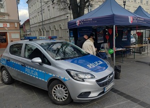 Na zdjęciu oznakowany radiowóz, w tle namiot koloru niebieskiego, gdzie widać policjantkę oraz kobietę, mężczyznę i dziecko.