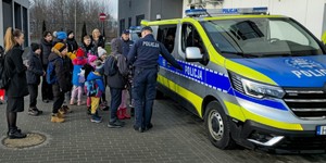 Grupa małych dzieci oraz trzy kobiety, a także dwaj umundurowani policjanci, którzy prezentują oznakowany radiowóz. W tle budynek komendy.