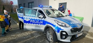 Dzieci oglądające oznakowany radiowóz. W tle policjant i budynek komendy.