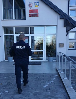 Umundurowany policjant stojący tyłem na tle wejścia do budynku szkoły. Nad szklanymi drzwiami jest Godło Polski, Herb oraz dwie tabliczki z nazwa szkoły.