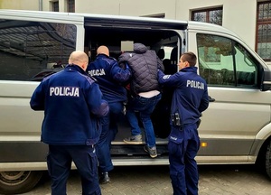 Na zdjęciu trzech umundurowanych policjantów, którzy wprowadzają do samochodu zatrzymanego mężczyznę, ubranego w niebieskie dżinsy i czarna kurtkę. W tle budynek