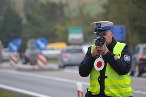 Umundurowany policjant ruchu drogowego trzymający w ręku policyjny miernik oraz tarcze sygnalizacyjną. W tle droga i samochody.