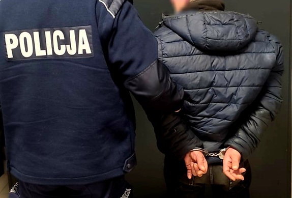 Na zdjęciu widać po lewej stronie umundurowanego policjanta , który trzyma zatrzymanego mężczyznę, który ma założone kajdanki ma ręce trzymane  z tyłu. Osoby są tyłem.