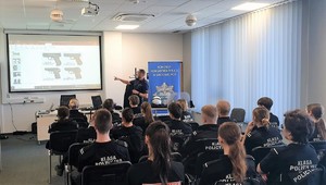 Grupa uczniów siedzących na krzesłach, osoby są tyłem. Ubrani w czarne koszulki z napisem klasa policyjna. Przed nimi policjant, który wskazuje na ekran, na którym są wyświetlane rodzaje broni.