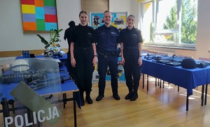 Umundurowany policjant, po prawej i po lewej stronie uczennice w szklonych mundurowych. W tle gazetka, na której są zdjęcia. Na stolikach widać poukładany sprzęt policyjny.