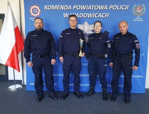 Na środku baneru z napisem Komenda Powiatowa Policji w Wadowicach stoi policjantka i policjant trzymający w ręku puchar.  Po prawej i po lewej stronie umundurowany policjant. Po lewej flaga Polski.