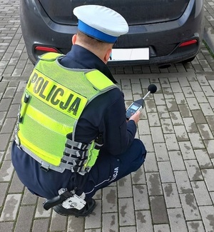 Umundurowany policjant ruchu drogowego w odblaskowej kamizelce z napisem Policja, który za pomocą sonometru sprawdza normy hałasu emitowane przez pojazd.