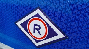 Na niebieskim tle logo ruchu drogowego. W białym rombie jest mniejszy romb koloru granatowego, w którym znajduje się białe koło z czerwoną otoczka. Wewnątrz koła jest litera R.