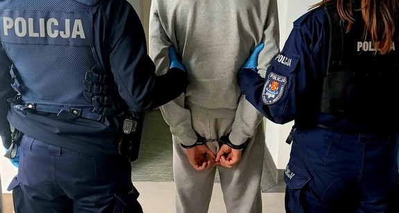 Na zdjęciu zatrzymany mężczyzna, w szarym dresie, który ma założone kajdanki na rękach trzymanych  z tyłu. Po lewej stronie policjant, po prawej stronie policjantka.