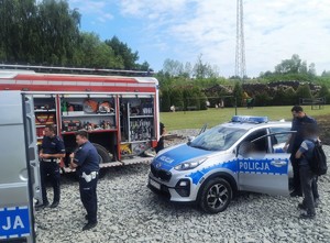 Na zdjęciu wóz strażacki, obok stoją dwaj policjanci. Po prawej stronie radiowóz, obok stojący policjant i mężczyzna.