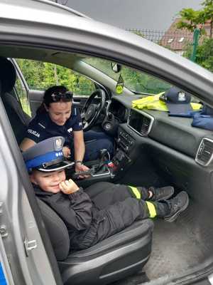 W radiowozie siedzi na miejscu kierowcy policjantka, a obok chłopiec, który ma złożoną policyjna czapkę.