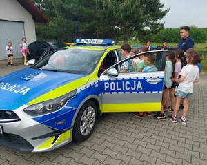 Na zdjęciu oznakowany radiowóz i grupa dzieci. Część z nich stoi wokół pojazdu, są też osoby wewnątrz. Po prawej stronie umundurowany policjant.