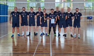 Reprezentacja wadowickiej komendy w piłce siatkowej. Dziesięciu mężczyzn w granatowych strojach sportowych, na szyi mają medale, Dwóch z nich ma zamazaną twarz. osoby stoją obok siebie na sali gimnastycznej.