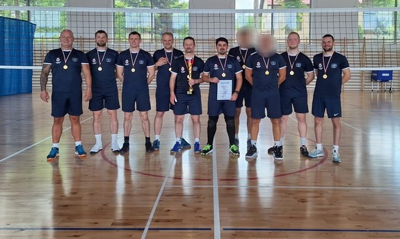 Reprezentacja wadowickiej komendy w piłce siatkowej. Dziesięciu mężczyzn w granatowych strojach sportowych, na szyi mają medale, Dwóch z nich ma zamazaną twarz. osoby stoją obok siebie na sali gimnastycznej.
