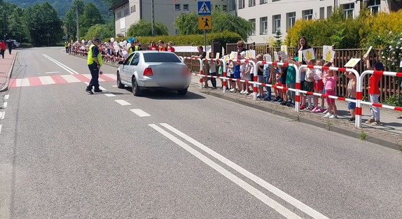 Na zdjęciu dzieci i osoby dorosłe stojące na chodniku przed szkołą z różnymi transparentami. Na drodze policjant kontroluje samochód.