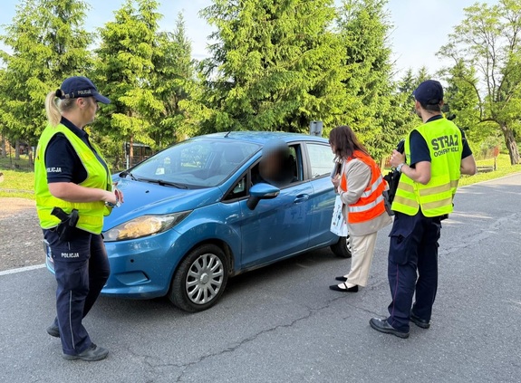 Na zdjęciu po lewej stronie policjantka, po prawej policjant i kobieta w pomarańczowej kamizelce odblaskowej i niebieski samochód osobowy.