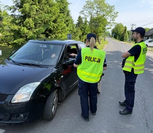 Na zdjęciu policjantka i policjant w odblaskowych kamizelkach z napisem Policja oraz czarny samochód osobowy.,