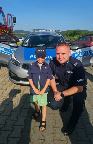 Na zdjęciu mały chłopczyk ubrany w koszulę i czapkę policyjną, obok umundurowany policjant. W tle oznakowany radiowóz i inne pojazdy.
