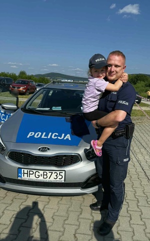 Umundurowany policjant, który trzyma na rękach małą dziewczynkę, która się do niego przytula. Dziewczynka ma założona policyjna czapkę. obok radiowóz, a w tle i inne pojazdy.