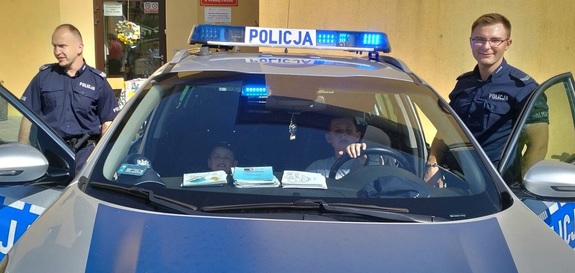 Na zdjęciu po prawej i po lewej stronie stoi umundurowany policjant . W radiowozie siedzi dwóch chłopców,  których jeden ma założona policyjną czapkę.