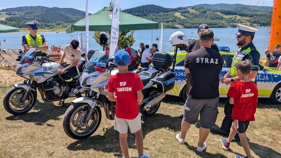 Na tle jeziora i namiotu widać dwa motocykle policyjne i radiowóz, a  także policjantów oraz grupę osób.