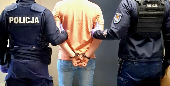 Zatrzymany mężczyzna, który ma założone kajdanki na rękach trzymanych z tyłu. Po prawej i po lewej stronie są umundurowani policjanci. Osoby są tyłem,