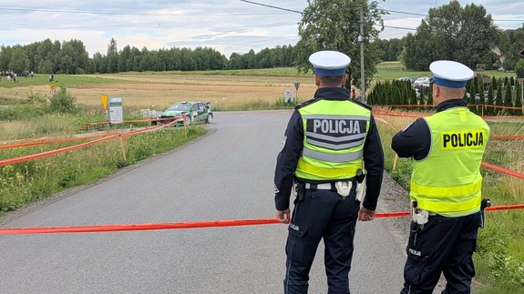 Dwóch umundurowanych policjantów ruchu drogowego zabezpieczających trasę rajdu. Są tyłem. Na drodze widać samochód rajdowy.