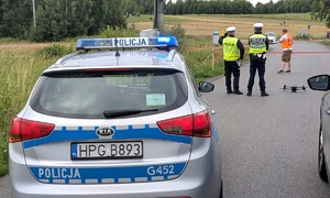 Po lewej radiowóz, po prawej dwóch policjantów ruchu drogowego i mężczyzna w pomarańczowej kamizelce odblaskowej. Osoby są tyłem. Na drodze dron. W tle samochód rajdowy.