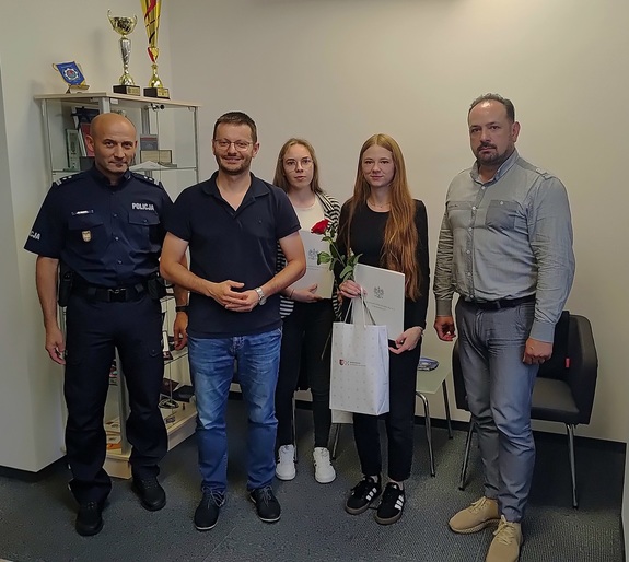 Od lewej umundurowany policjant, dalej mężczyzna w stroju cywilnym, dwie kobiety trzymające w rękach różę torbę papierową oraz po prawej mężczyzna w stroju cywilnym.
