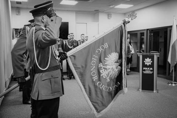 Na zdjęciu widać poczet sztandarowy, trzech policjantów, z których policjant stojący w środku trzyma sztandar wadowickiej komendy. W tle stoją policjanci.
