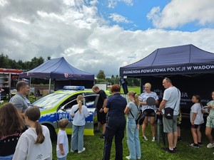 policjanci na pikniku rodzinnym, w tle oznakowany radiowóz, stoisko policyjne, osoby uczestniczace w pikniku ogladają policyjne wyposażenie