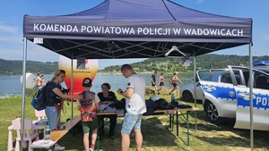 stoisko policyjne, w tle jezioro Mucharskie, radiowóz, osoby wypczywające zainteresowane stoiskiem
