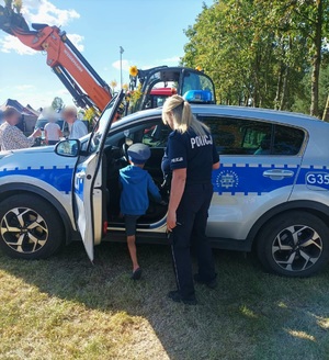 Chłopczyk w policyjnej czapce wchodzący do radiowozu. Obok stoi policjantka.