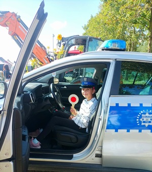 Dziewczynka siedząca na miejscu kierowcy w radiowozie. Na głowie ma założona policyjną czapkę, a w rękach trzyma tarcze sygnalizacyjną.