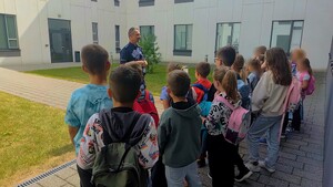 Umundurowany policjant, przed którym stoi grupa dzieci. W tle budynek komendy.