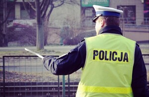 Policjant ruchu drogowego stojący tyłem, ma założona kamizelkę odblaskową z napisem Policja, w lewej ręce trzyma tarcze sygnalizacyjną
