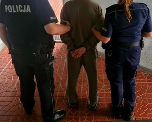 Po lewej stronie umundurowany policjant , po prawej policjantka, a po środku zatrzymany mężczyzna, który ma założone kajdanki na ręce trzymane z tyłu.