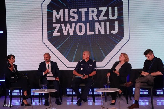 Na środku siedzi policjant, po bokach dwóch mężczyzn i dwie kobiety w strojach cywilnych. Nad nimi baner z napisem Mistrzu zwolnij