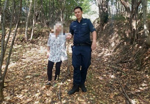 Umundurowany policjant prowadzący pod rękę starszą kobietę. Osoby są w lesie.