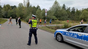 Policjant ruchu drogowego stojący na drodze, po prawej stronie radiowóz. W tle uczestnicy biegu.