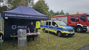 Po lewej wóz strażacki i radiowóz, a po prawej namiot, gdzie stoi policjant a na stoliku poukładane są ulotki i elementy umundurowania policyjnego.