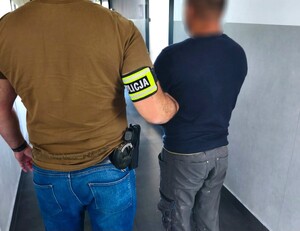 Na zdjęciu po lewej stronie policjant w stroju cywilnym z bronią na pasku i opaska z napisem Policja na prawej ręce, który trzyma zatrzymanego mężczyznę.