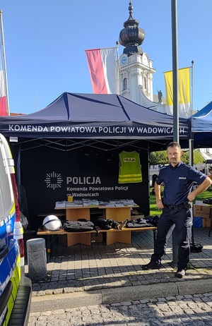 Namiot z napisem Komenda Powiatowa Policji w Wadowicach, w którym na stoliku poukładany jest sprzęt policyjny i materiały profilaktyczne. Po prawej stronie policjant.