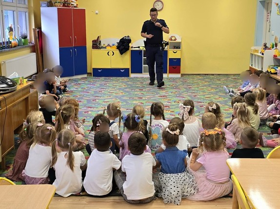 Grupa dzieci siedzących na dywanie, przed nimi policjant.