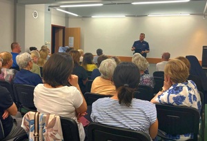 Grupa osób siedzących na krzesłach, osoby są tyłem. Przed nimi umundurowany policjant.