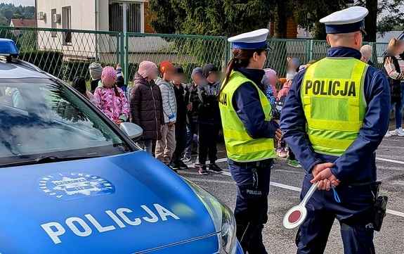 Umundurowana policjantka i policjant ruchu drogowego, po lewej stronie radiowóz, w tle grupa dzieci.