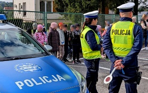 Umundurowana policjantka i policjant ruchu drogowego, po lewej stronie radiowóz, w tle grupa dzieci.